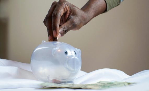 Close-up of a person's hand placing coins into a transparent piggy bank to save money.
